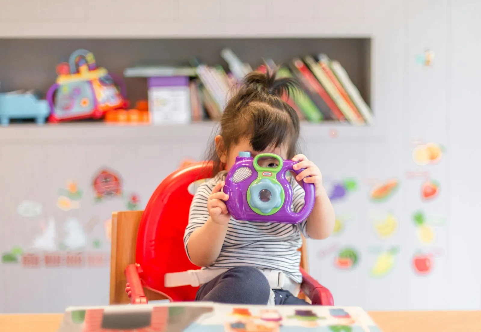 Niña pequeña sentada en silla roja sosteniendo una cámara de juguete púrpura, en un entorno de aprendizaje con juguetes y libros de fondo, representando la integración de terapia y educación en Chroma Early Start.