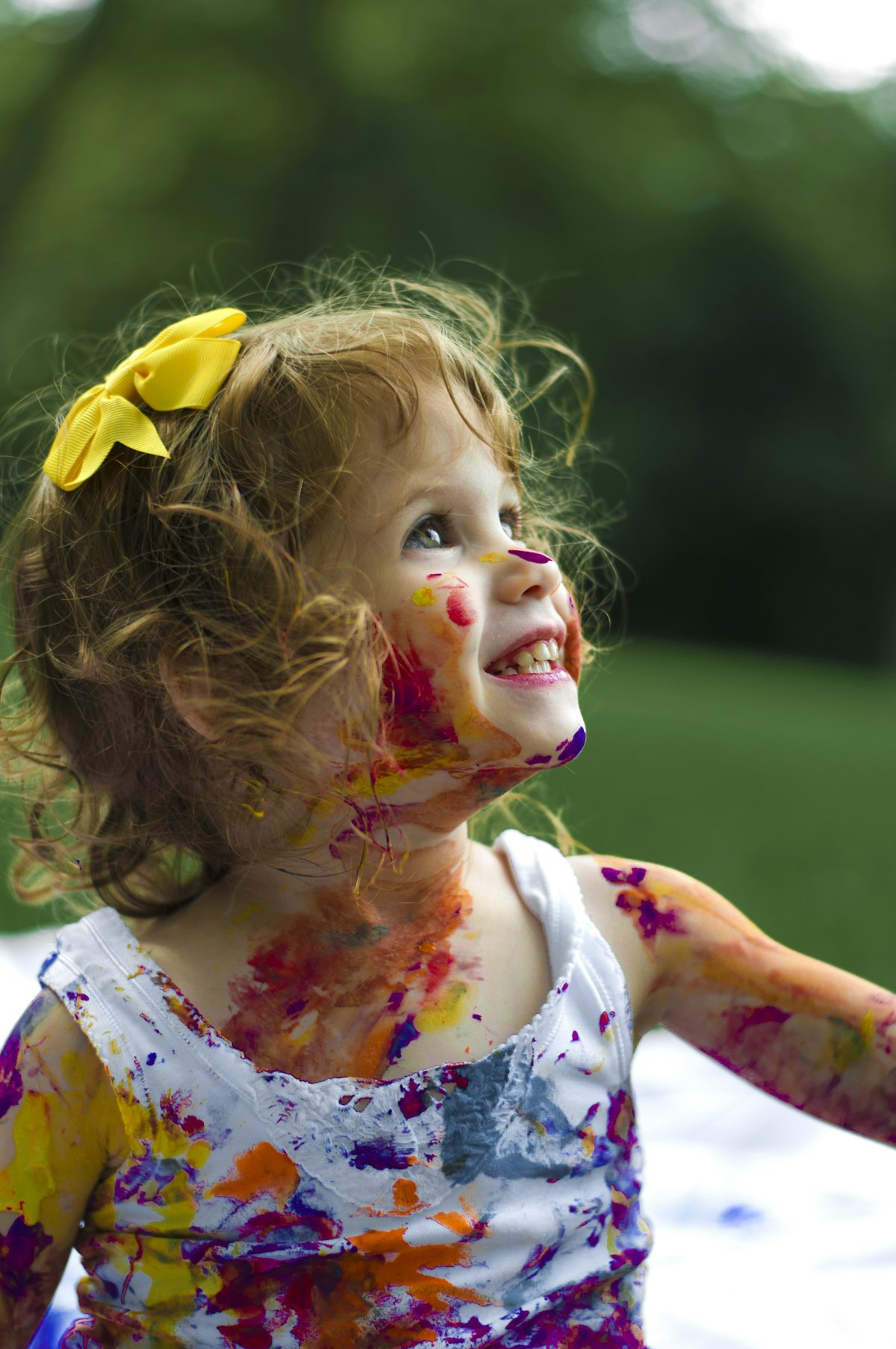 Niña sonriente con cabello rizado y moño amarillo, cubierta de pintura colorida en la cara y el cuerpo, disfrutando de una experiencia creativa en un entorno al aire libre, reflejando un enfoque inclusivo y lúdico en el desarrollo infantil.