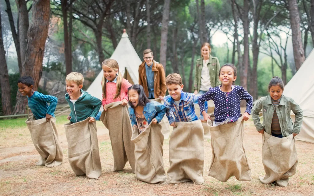 Children participating in a sack race outdoors, laughing and engaging in playful competition, with tents and trees in the background, emphasizing social readiness and active play in Chroma Early Learning Academy's Pre-K program.