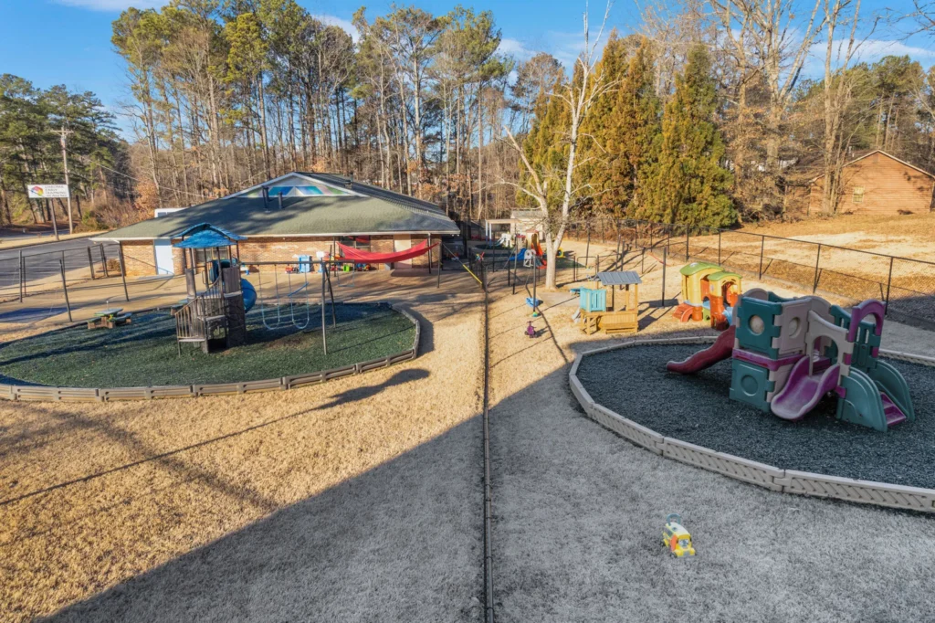 Playground area at Chroma Early Learning Academy's Marietta Campus, featuring swings, slides, and play structures, surrounded by a secure fence and trees, emphasizing a safe and engaging environment for children.