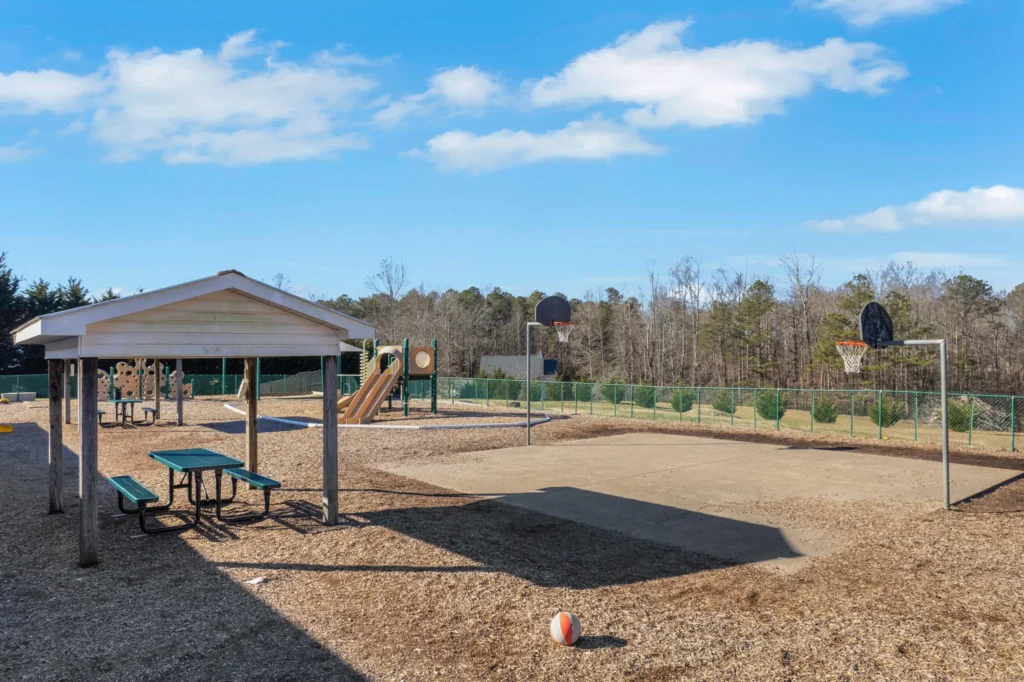 Playground area at Cherokee Academy featuring a basketball court, playground equipment, picnic tables, and a blue sky, emphasizing a safe and engaging environment for children.