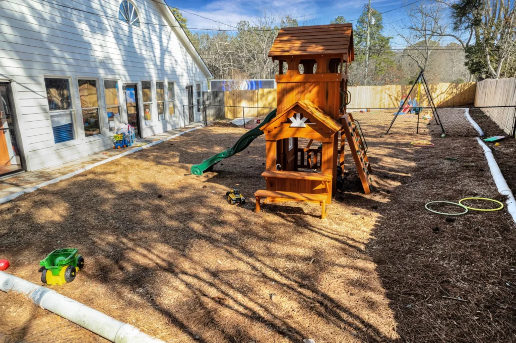 Playground area at Chroma Early Learning Academy Ellenwood, featuring a wooden play structure with slide, swings, and toys on a mulch surface, surrounded by a fenced yard and classroom windows.