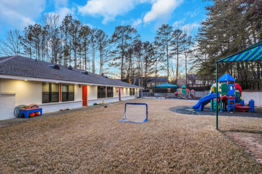 Outdoor play area at Chroma Early Learning Academy in Johns Creek, featuring colorful playground equipment, grassy space, and a secure building with large windows.