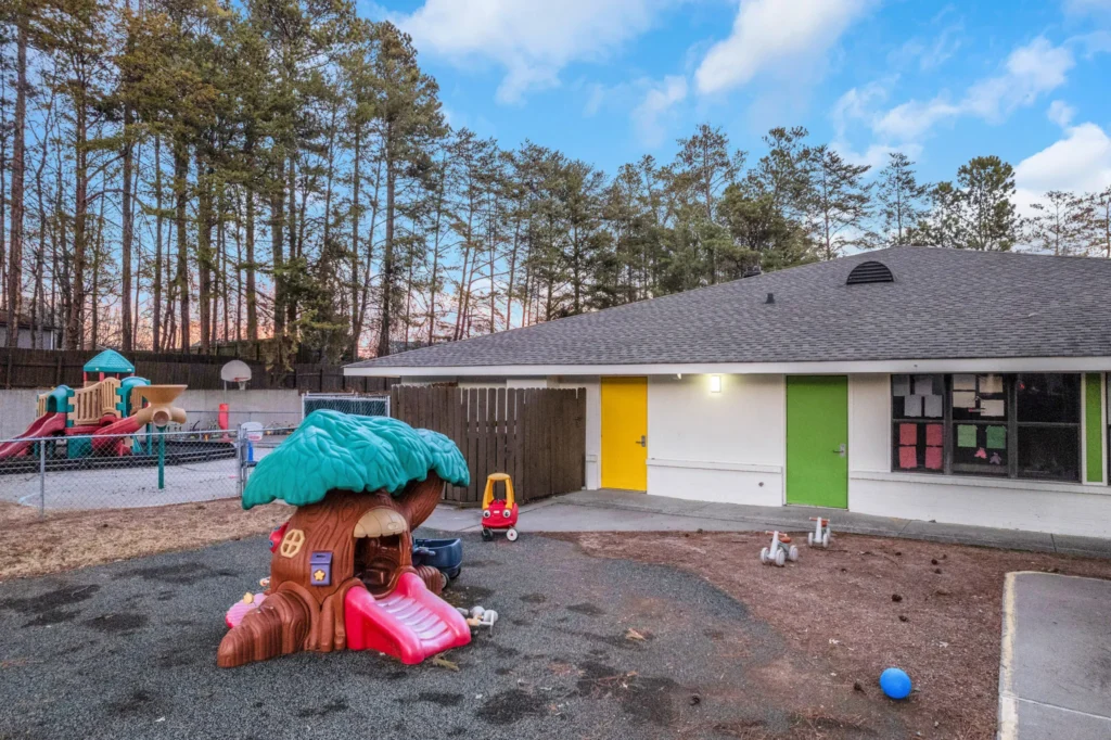 Playground area at Chroma Early Learning Academy featuring a colorful treehouse slide, adjacent play structures, and a fenced outdoor space surrounded by trees, emphasizing a safe and engaging environment for children.