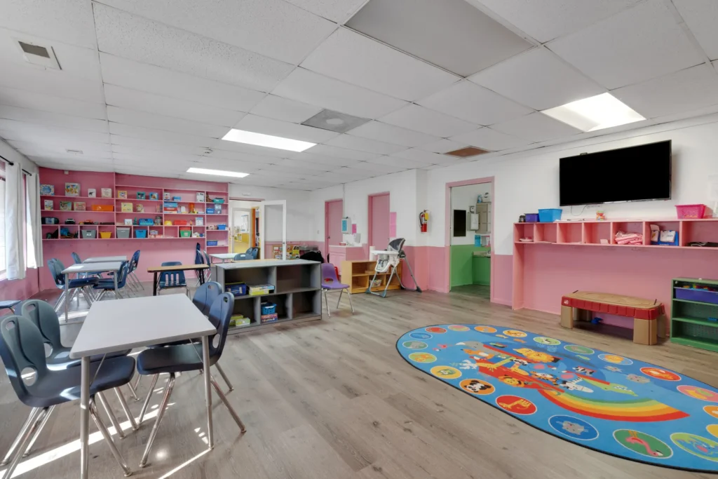 Classroom interior at Chroma Early Learning Academy's Tramore Campus, featuring colorful shelves, child-sized tables and chairs, a play rug, and a wall-mounted TV, designed for early education and childcare.