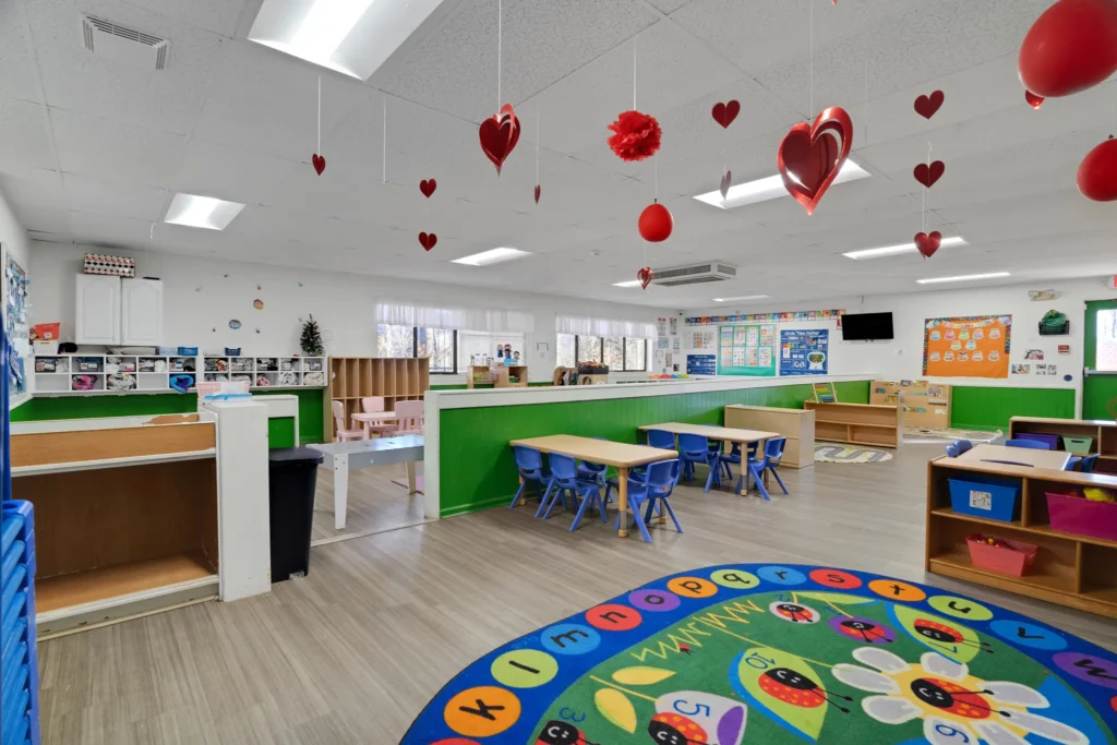 Brightly colored classroom at Chroma Early Learning Academy featuring green walls, child-sized tables and chairs, a circular alphabet rug, and festive hanging decorations, emphasizing a nurturing and engaging environment for early childhood education.