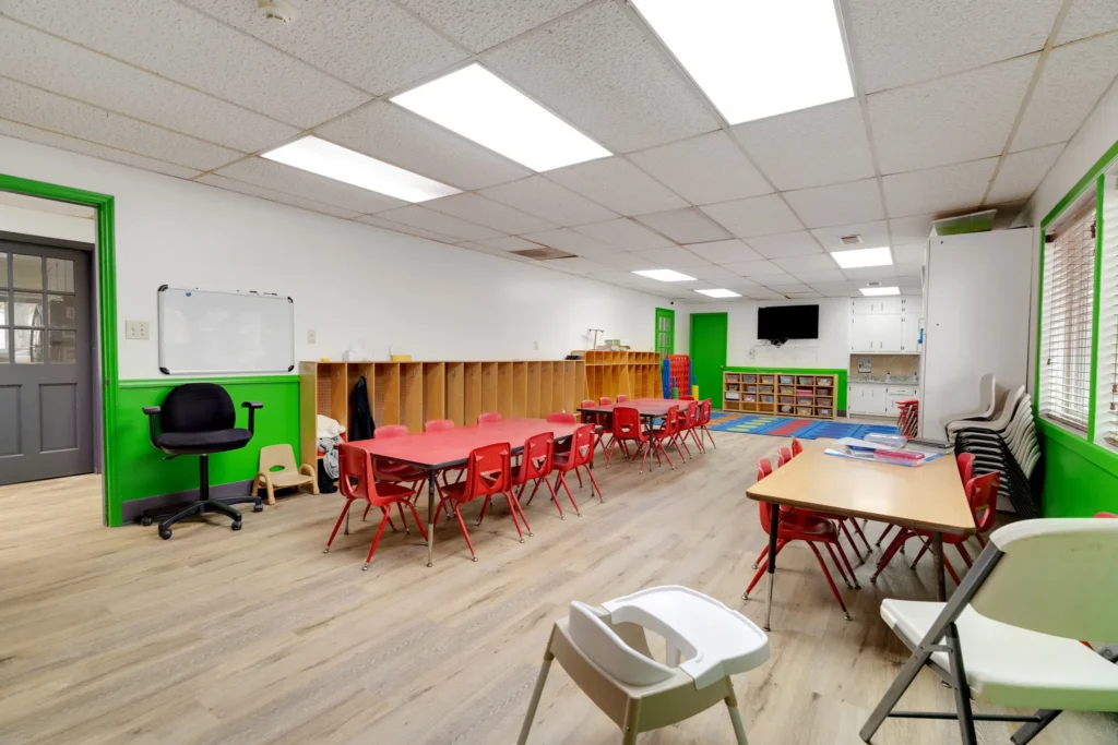 Early learning classroom at Chroma Early Learning Academy featuring colorful red chairs, a whiteboard, storage shelves, and a vibrant green accent wall, designed for nurturing children's growth and development.