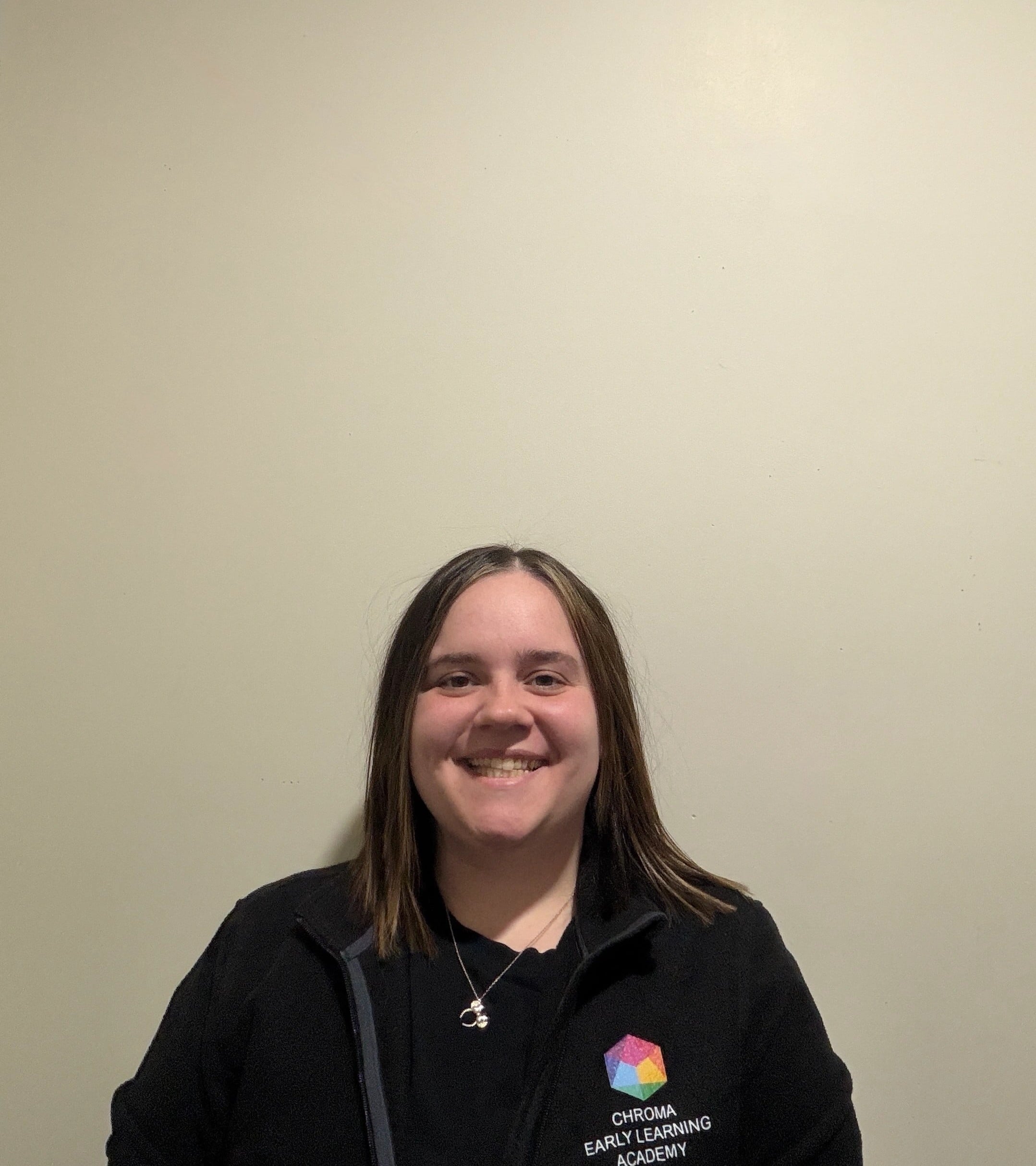 Directora de Chroma Early Learning Academy sonriendo, vestida con chaqueta negra con logotipo, fondo neutro.