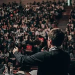 Hombre hablando frente a una multitud en un evento, destacando la importancia de la educación y el aprendizaje en la comunidad.