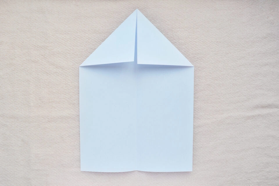 A folded sheet of light blue paper with upper corners bent toward the center, illustrating the initial steps of creating a paper airplane for early learning STEM activities.