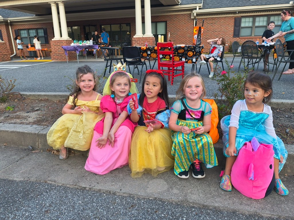 Five young girls in colorful princess costumes, smiling and sitting together on a curb during a community event at Chroma Early Learning Academy.