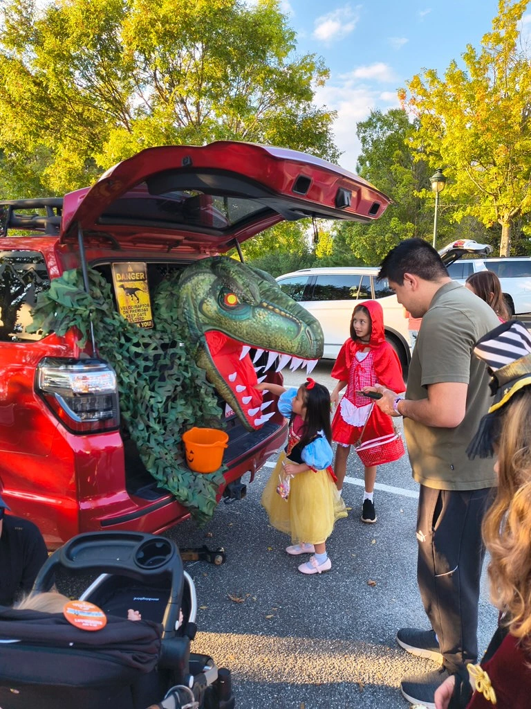 Children in costumes engaging at a trunk-or-treat event, featuring a decorated red vehicle with a dinosaur theme, surrounded by festive autumn foliage.