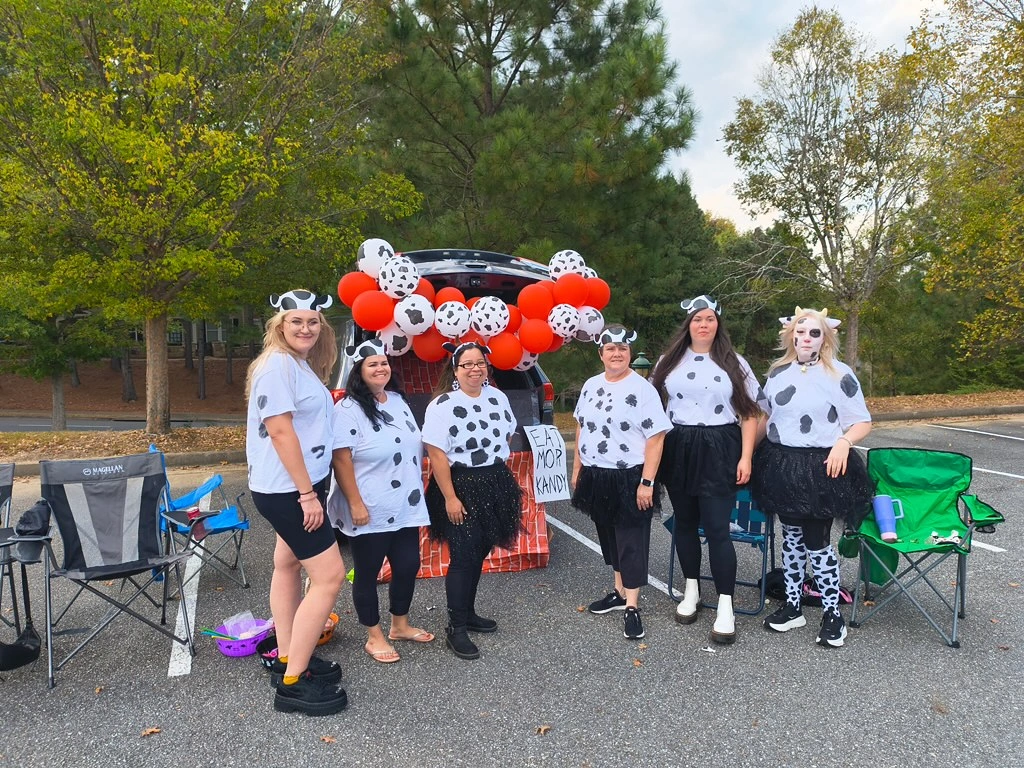 Group of adults dressed in cow-themed costumes with black and white spots, gathered at a festive event featuring balloons and a sign reading "EAT MOR KANDY," celebrating community and family engagement at Chroma Early Learning Academy.