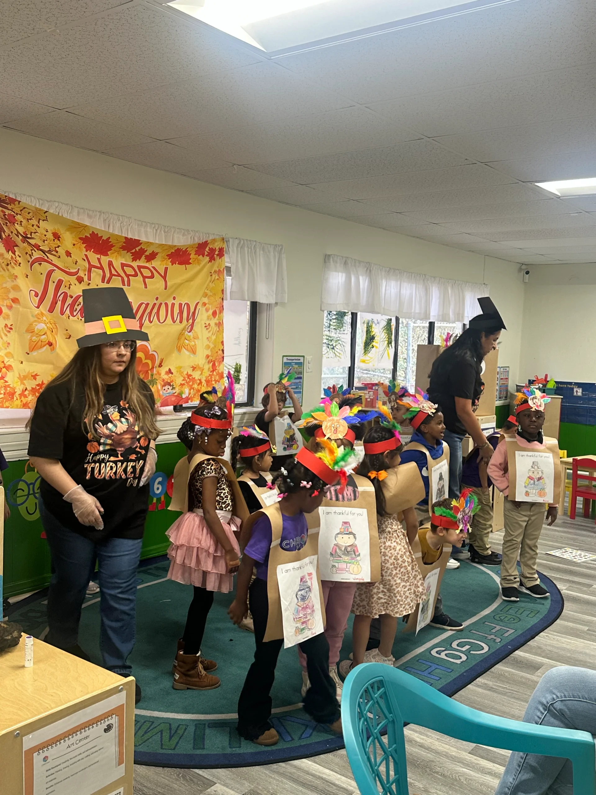 Children in Thanksgiving costumes performing at Chroma Early Learning Academy event, featuring colorful hats and shirts, with a festive backdrop reading "Happy Thanksgiving."